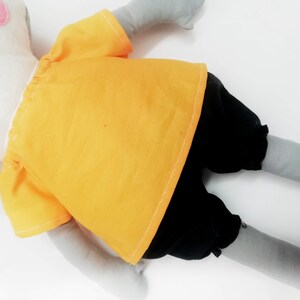 Puede incluir: Un juguete de peluche hecho a mano en posici&oacute;n acostada, con una cabeza blanca con detalles rosas y extremidades grises. Lleva una camisa amarilla y pantalones cortos negros. El juguete es para ni&ntilde;os.