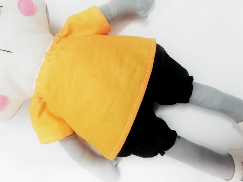 Puede incluir: Un juguete de peluche hecho a mano en posici&oacute;n acostada, con una cabeza blanca con detalles rosas y extremidades grises. Lleva una camisa amarilla y pantalones cortos negros. El juguete es para ni&ntilde;os.