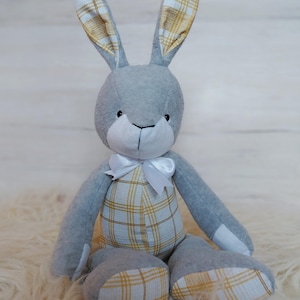May include: A grey plush rabbit toy with a white bow. The bunny's ears and plaid overalls have a yellow and white pattern. The toy is sitting on a fluffy, white surface.