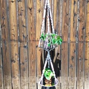 May include: A white macrame plant hanger with two circular wooden shelves. The hanger is hanging from a wooden fence and has two potted plants.