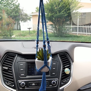 May include: A blue macrame plant hanger with a small white pot and a green succulent plant inside. The hanger is hanging from the rearview mirror of a car.