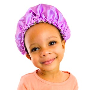May include: A young girl with dark brown skin is wearing a purple satin sleep cap. She is smiling and looking at the camera.