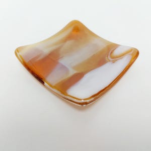 May include: A diamond-shaped glass dish with a warm color palette of orange, white, and brown. The dish has a glossy finish and a slightly raised edge. The design features a marbled effect, with the colors blending together in a unique pattern.