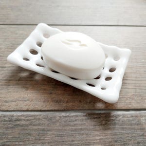 May include: A white ceramic soap dish holding a white bar of soap. The dish is rectangular with a wavy edge and circular drainage holes. The soap has a curved design on its surface. The soap dish is made of ceramic.