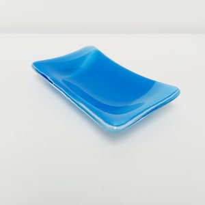 May include: A small, rectangular, blue glass dish with a white rim. The dish is slightly curved and has a glossy finish.