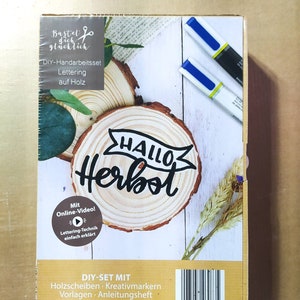 Op de afbeelding: Een DIY-knutselset voor het letteren op hout. De set bevat houten schijven, stiften, sjablonen en instructies. De afbeelding toont een houten schijf met de woorden "Hallo Herbst" erop.