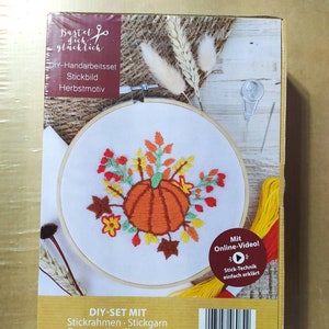 Op de afbeelding: Een DIY-borduurset met een herfstthema met een pompoen, bladeren en bloemen. De set bevat een houten borduurring, borduurgaren, stof, naalden en instructies. De doos heeft de tekst "Bastel dich glücklich" en "DIY-Handarbeitsset Stickbild Herbstmotiv".