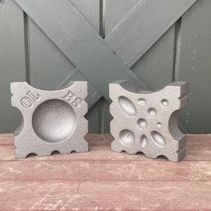 May include: Two grey metal blocks with distinct designs. One block has a central circular indentation and the letters "OL BS" etched on the surface. The other block features a series of oval and circular indentations. Both blocks are square with curved edges.