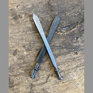 Può includere: Due oggetti in metallo forgiati a mano a forma di spade in miniatura con punte acuminate e manici arrotondati con pomolo. Gli elementi in ferro grigio scuro sono posizionati a forma di X, con una finitura metallica testurizzata e martellata.