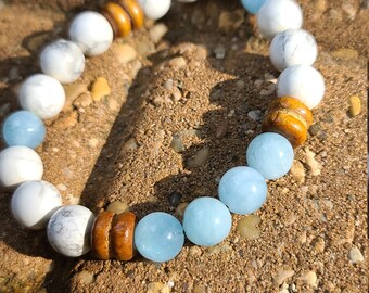 May include: A bracelet made with white and light blue beads and brown wooden beads. The bracelet is lying on a brown surface.