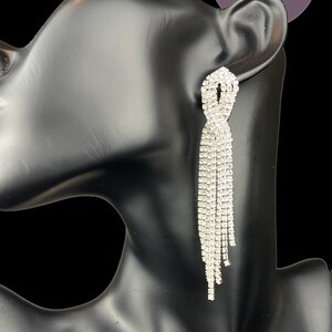 May include: A pair of dazzling, silver-colored rhinestone earrings. The earrings feature a knotted design at the top, with multiple strands of rhinestones cascading downwards. The earrings are displayed on a black mannequin. The text "Sparkle Competition Jewelry" is in the background.