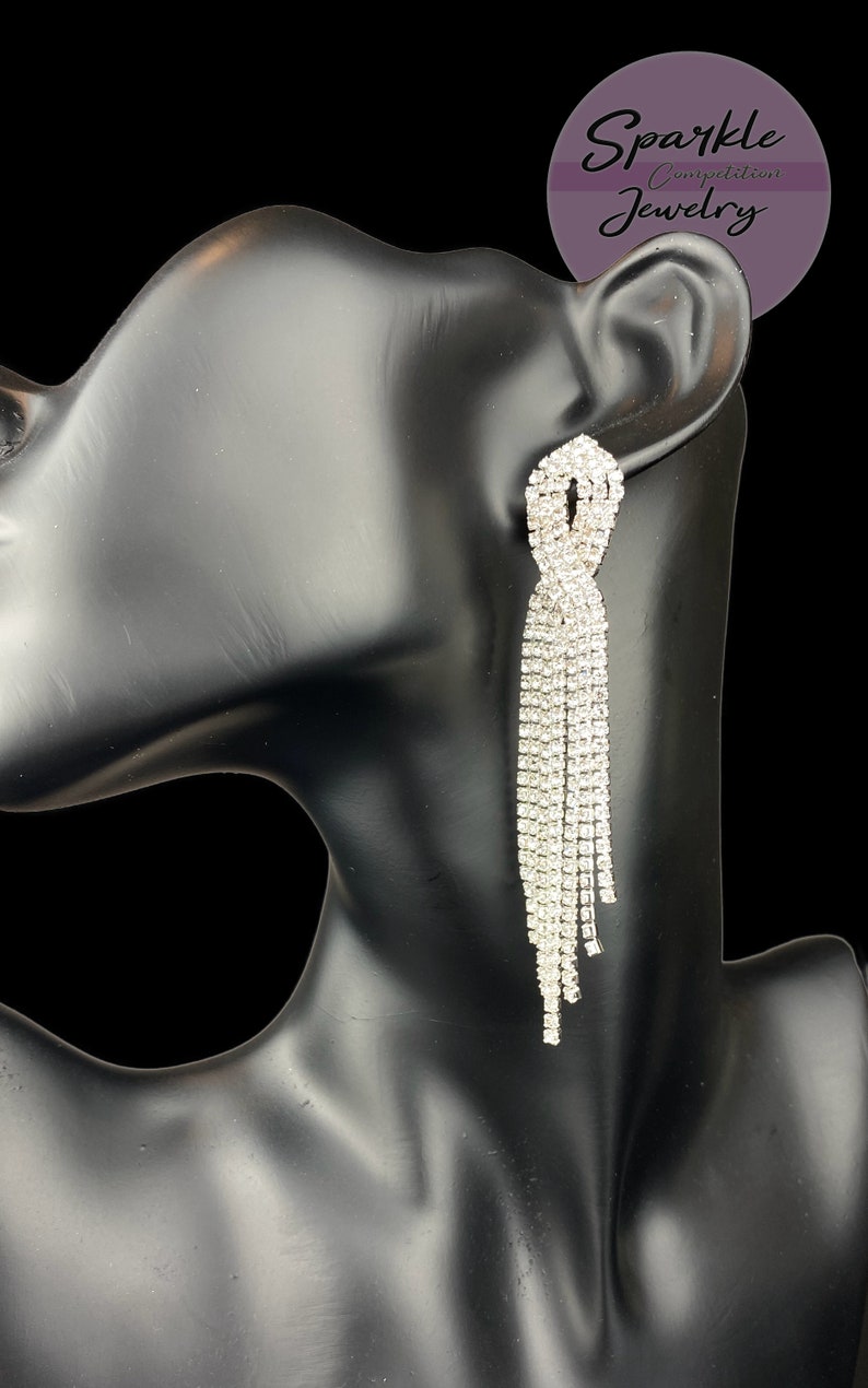 May include: A pair of dazzling, silver-colored rhinestone earrings. The earrings feature a knotted design at the top, with multiple strands of rhinestones cascading downwards. The earrings are displayed on a black mannequin. The text "Sparkle Competition Jewelry" is in the background.