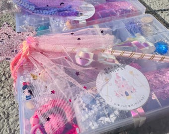 May include: A clear plastic storage box filled with pink and purple princess-themed party supplies. The box includes a pink tulle skirt, a pink wand, a pink and gold crown, and a pink and gold castle-shaped sticker that says "Happy Birthday".