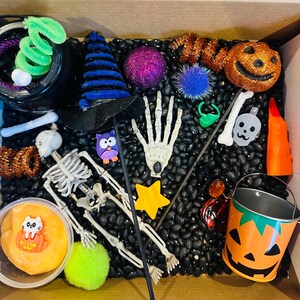 May include: A black sensory bin filled with black beans and Halloween themed toys, including a skeleton, a witch's hat, a pumpkin, an owl, a hand, a star, a cauldron, and a bucket.