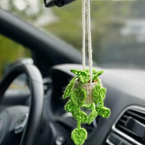 May include: A handmade crochet car charm featuring a small, cream-colored pot holding a vibrant green plant with heart-shaped leaves. The charm hangs from a white string, adding a touch of nature to the car's interior.