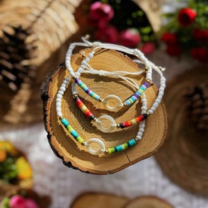 Könnte beinhalten: Drei handgefertigte Armbänder mit weißen und bunten Perlen. Jedes Armband hat ein weißes, oval geformtes Mittelstück und goldfarbene Akzente. Die Armbänder werden auf einer Holzoberfläche präsentiert und zeigen einen Bohemian-Stil.