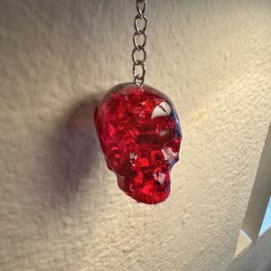 May include: A red, translucent skull-shaped keychain. The keychain is made of a shiny, possibly resin material, with a silver chain and keyring. The skull has visible details and a textured appearance.