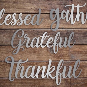 May include: Silver metal wall decor with the words "Blessed gather grateful thankful" in a cursive font on a wood background.