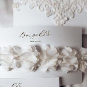 A collection of delicate bridal accessories is displayed on elegant white cards. The accessories include a lace garter, a floral chiffon garter, and a pearl-embellished garter, all in soft ivory tones. The presentation exudes a romantic and sophisticated bridal aesthetic.