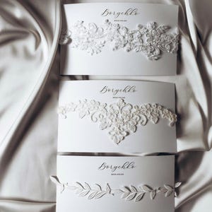 A collection of delicate bridal accessories is displayed on elegant white cards. The accessories include a lace garter, a floral chiffon garter, and a pearl-embellished garter, all in soft ivory tones. The presentation exudes a romantic and sophisticated bridal aesthetic.