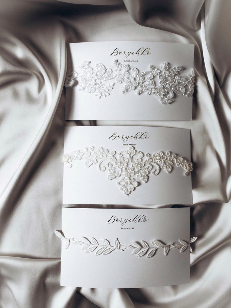 A collection of delicate bridal accessories is displayed on elegant white cards. The accessories include a lace garter, a floral chiffon garter, and a pearl-embellished garter, all in soft ivory tones. The presentation exudes a romantic and sophisticated bridal aesthetic.