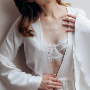 May include: A white satin robe with long, wide sleeves and a matching tie belt, worn over a white lace bralette featuring intricate floral patterns and scalloped edges along the neckline and underwire, creating a layered and textured appearance.