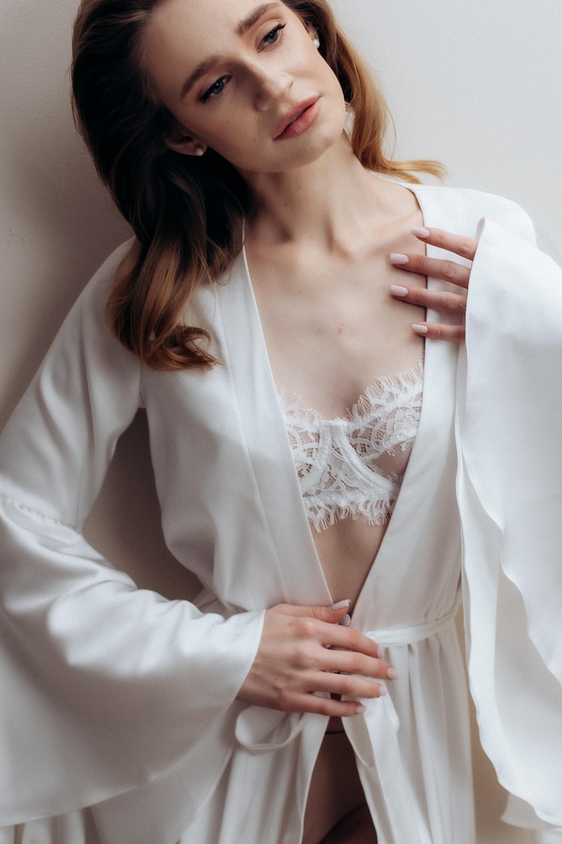 May include: A white satin robe with long, wide sleeves and a matching tie belt, worn over a white lace bralette featuring intricate floral patterns and scalloped edges along the neckline and underwire, creating a layered and textured appearance.