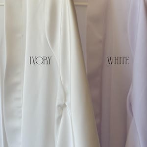 May include: Two satin robes hanging side by side. The robe on the left is ivory with the word IVORY printed in black. The robe on the right is white with the word WHITE printed in black. Both robes feature a smooth finish and open front design.