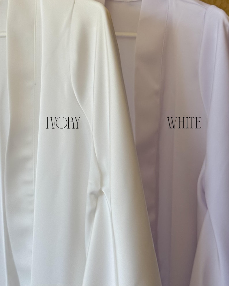May include: Two satin robes hanging side by side. The robe on the left is ivory with the word IVORY printed in black. The robe on the right is white with the word WHITE printed in black. Both robes feature a smooth finish and open front design.