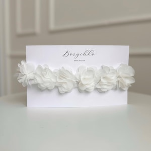 A delicate bridal garter from is displayed on a white card. It features soft ivory chiffon flowers arranged in a row, creating a romantic and elegant look. The garter is designed for brides seeking a timeless and feminine accessory for their special day.