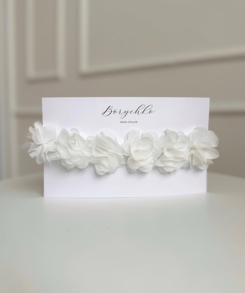 A delicate bridal garter from is displayed on a white card. It features soft ivory chiffon flowers arranged in a row, creating a romantic and elegant look. The garter is designed for brides seeking a timeless and feminine accessory for their special day.