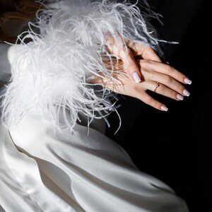 May include: A white satin dress with a feathered cuff. A hand with a ring on the ring finger is visible. The nails are painted with a light pink color. The background is dark.