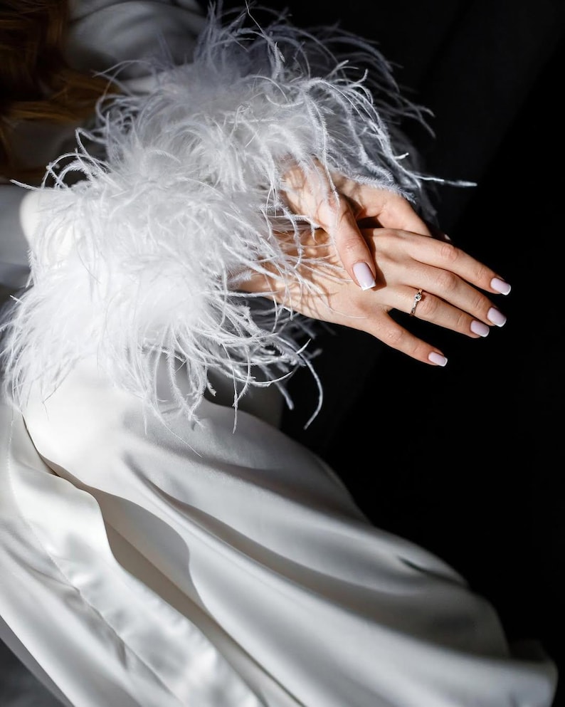 May include: A white satin dress with a feathered cuff. A hand with a ring on the ring finger is visible. The nails are painted with a light pink color. The background is dark.