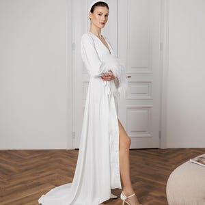 May include: A white satin robe with a long train and feather trim on the sleeves. The robe is open in the front, revealing a high leg slit. The model is wearing a white headband and white heels.