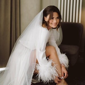 May include: A white satin bridal robe with long sleeves and feather trim on the cuffs. The bride is wearing a sheer veil and silver heels. The robe is knee-length and the bride is sitting in a chair.