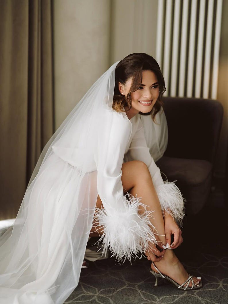 May include: A white satin bridal robe with long sleeves and feather trim on the cuffs. The bride is wearing a sheer veil and silver heels. The robe is knee-length and the bride is sitting in a chair.