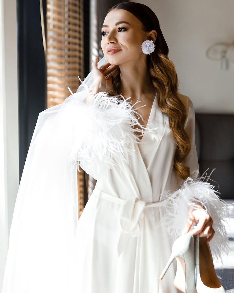May include: A white satin robe with feather trim on the sleeves and a matching belt. The robe is paired with white high-heeled shoes. The woman is wearing white flower earrings.