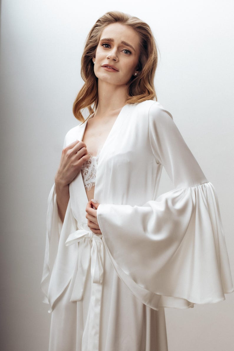May include: A white satin robe featuring long, wide bell sleeves and a matching tie belt at the waist. The robe has a deep V-neckline that reveals a white lace camisole underneath, creating a layered look with smooth, flowing fabric.