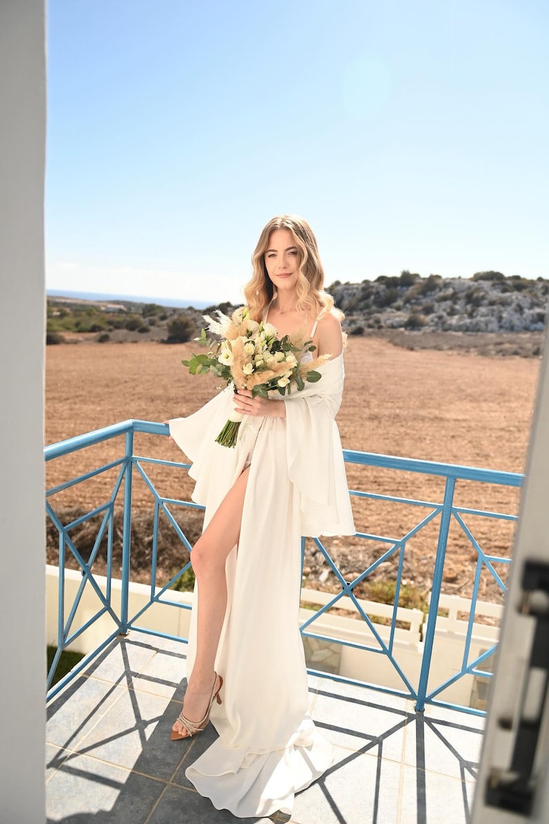 May include: A floor-length white dress with wide sleeves and a high leg slit, paired with strappy tan heels. The garment is worn with a bouquet of white roses, greenery, and dried pampas grass. The outfit features a simple neckline and flowing fabric.