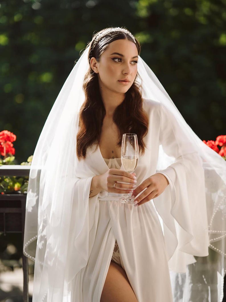 May include: A white satin robe featuring long, flared bell sleeves and a tie waist, paired with a sheer, floor-length veil adorned with delicate pearl-like embellishments along the edges and a matching pearl-accented headband.