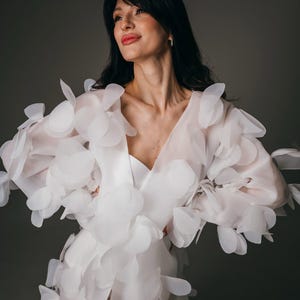 May include: A white wedding dress with a V-neckline and a slit. The dress features large, petal-like embellishments on the sleeves and bodice. The model is wearing the dress in front of a gray background.