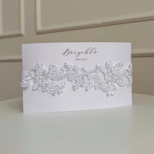 May include: A white lace garter with a white card that says 'Borychko Bride Atelier'.