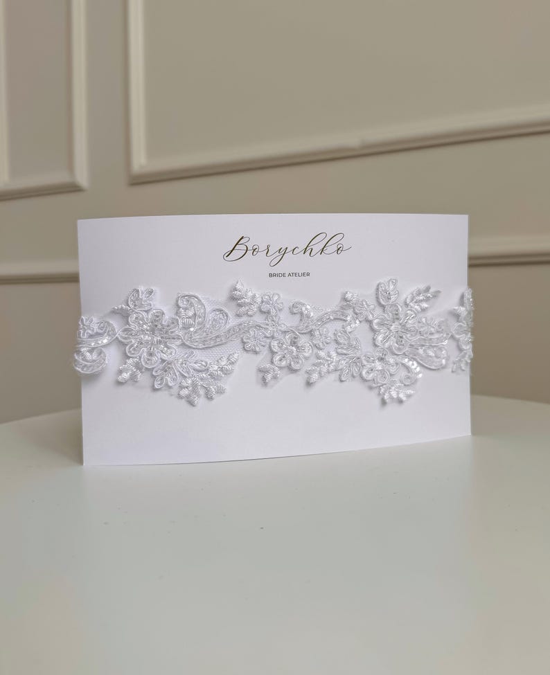 May include: A white lace garter with a white card that says 'Borychko Bride Atelier'.