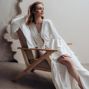 May include: A floor-length white robe featuring long, wide bell sleeves and a tie-waist closure. The garment is crafted from a smooth, satin-like fabric with a deep V-neckline and a high side slit that extends from the thigh to the hemline.