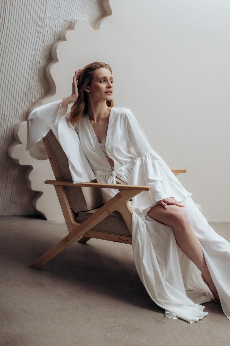 May include: A floor-length white robe featuring long, wide bell sleeves and a tie-waist closure. The garment is crafted from a smooth, satin-like fabric with a deep V-neckline and a high side slit that extends from the thigh to the hemline.