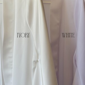 May include: Close-up of two robes, one ivory and one white, hanging on a white hanger. The word "IVORY" is printed in a serif font on the ivory robe, and the word "WHITE" is printed on the white robe. The robes appear to be made of a smooth, silky material.
