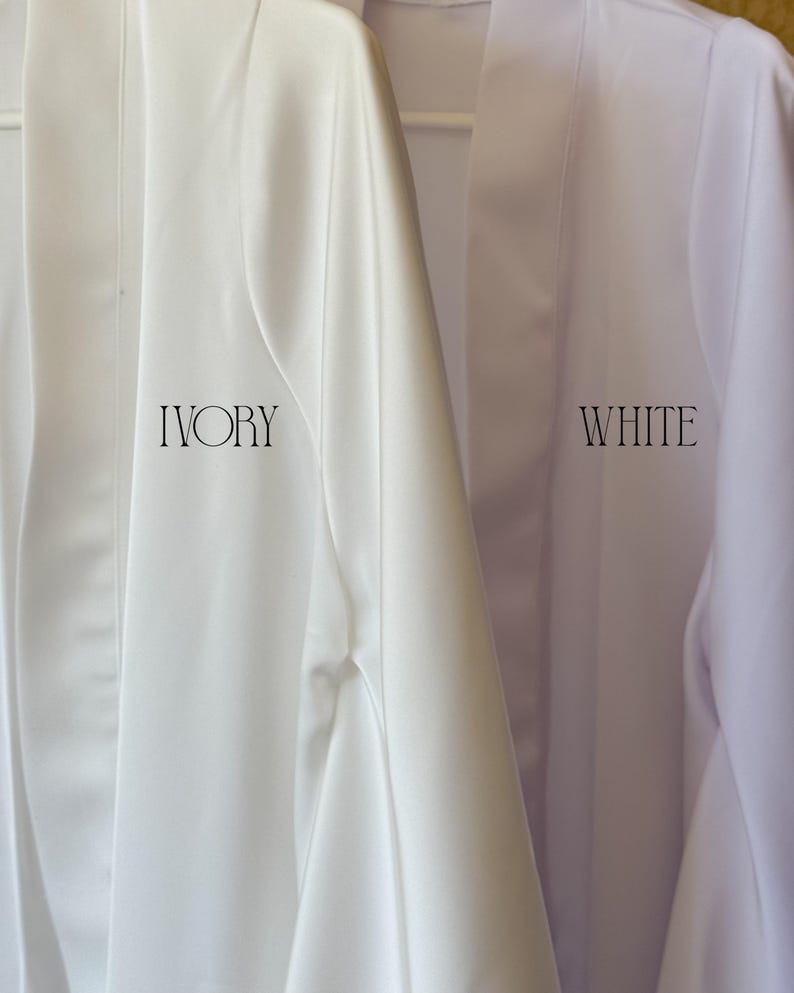 May include: Close-up of two robes, one ivory and one white, hanging on a white hanger. The word "IVORY" is printed in a serif font on the ivory robe, and the word "WHITE" is printed on the white robe. The robes appear to be made of a smooth, silky material.