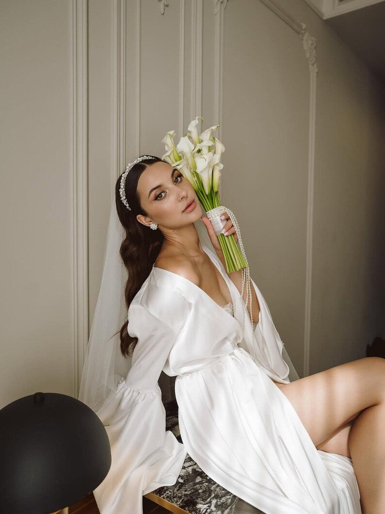 May include: A white satin robe with long bell sleeves, paired with a sheer veil and a pearl-embellished headband. The ensemble includes a bouquet of white calla lilies wrapped in a pearl-beaded strand, complemented by matching pearl stud earrings.