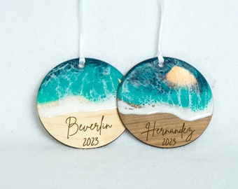 May include: Two round wooden ornaments with a beach scene design. The top half features turquoise and white resin waves, and the bottom half is a light wood beach. One ornament says "Beverlin 2023" and the other says "Hernandez 2023".