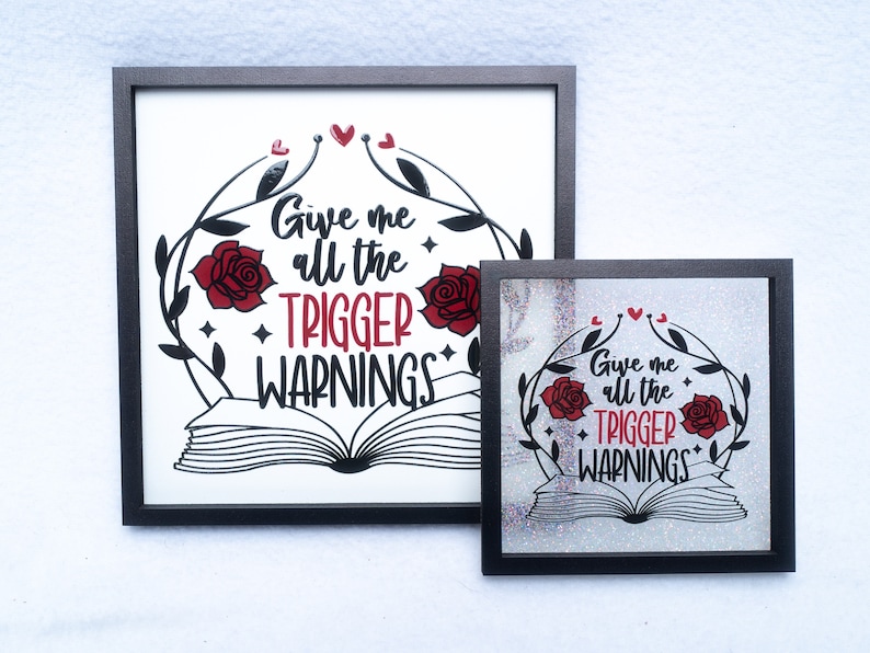 May include: Two framed prints with a black frame and a white background. The print features a black and white illustration of an open book with red roses and vines. The text on the print reads "Give me all the trigger warnings".